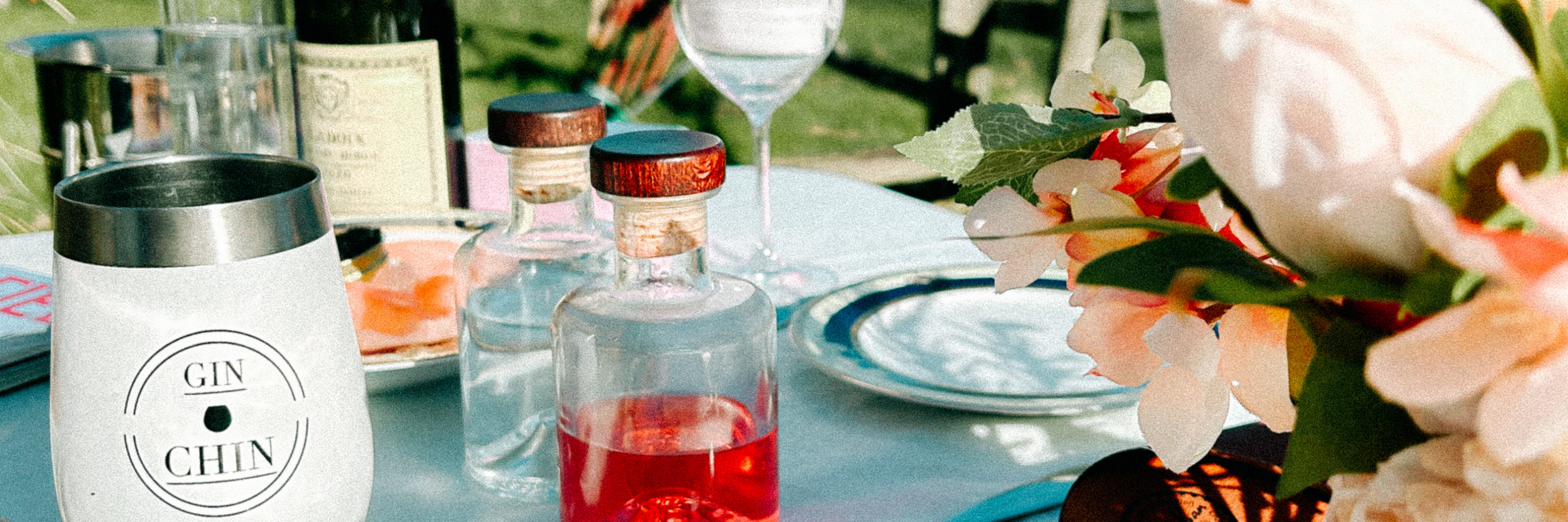 Table setting with artisan gin and tonic glasses, bottles, and flowers outdoors.