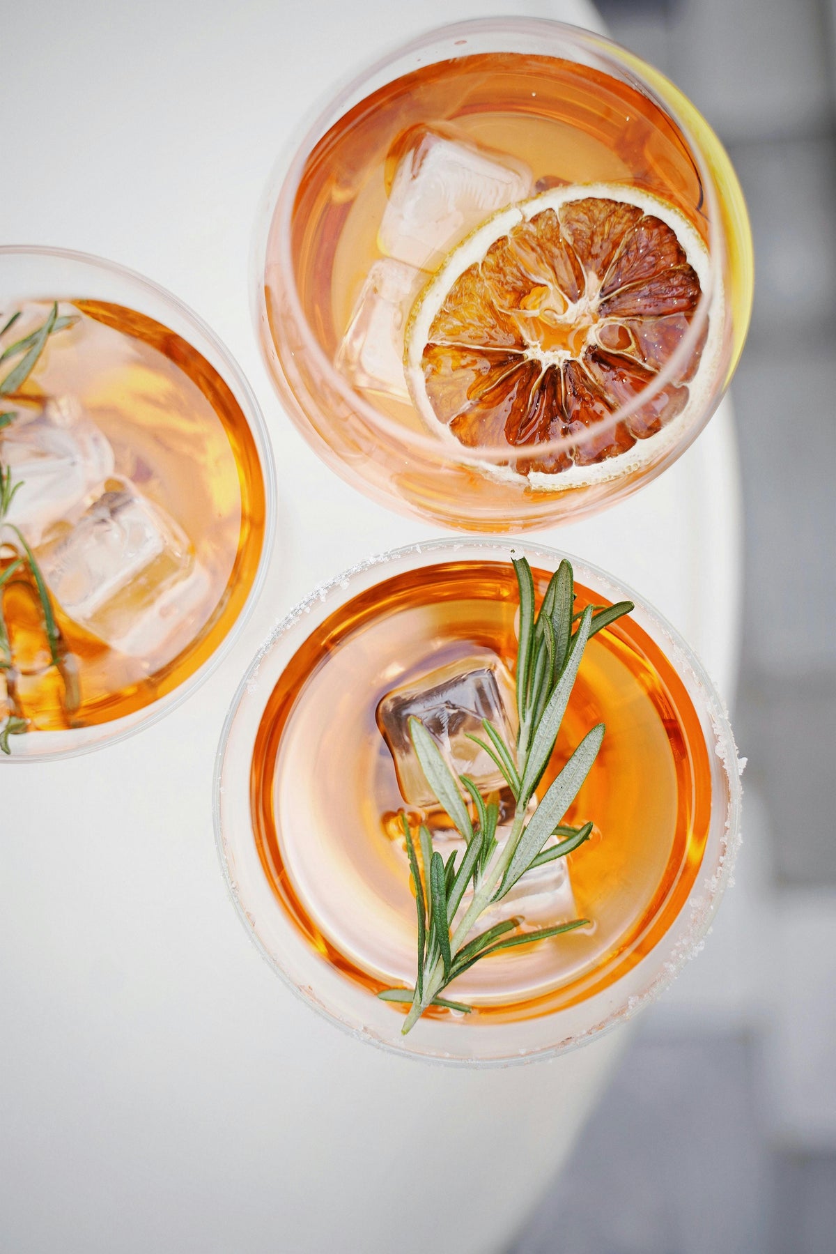 Three cocktails with ice cubes, orange slices, and rosemary on a white surface.