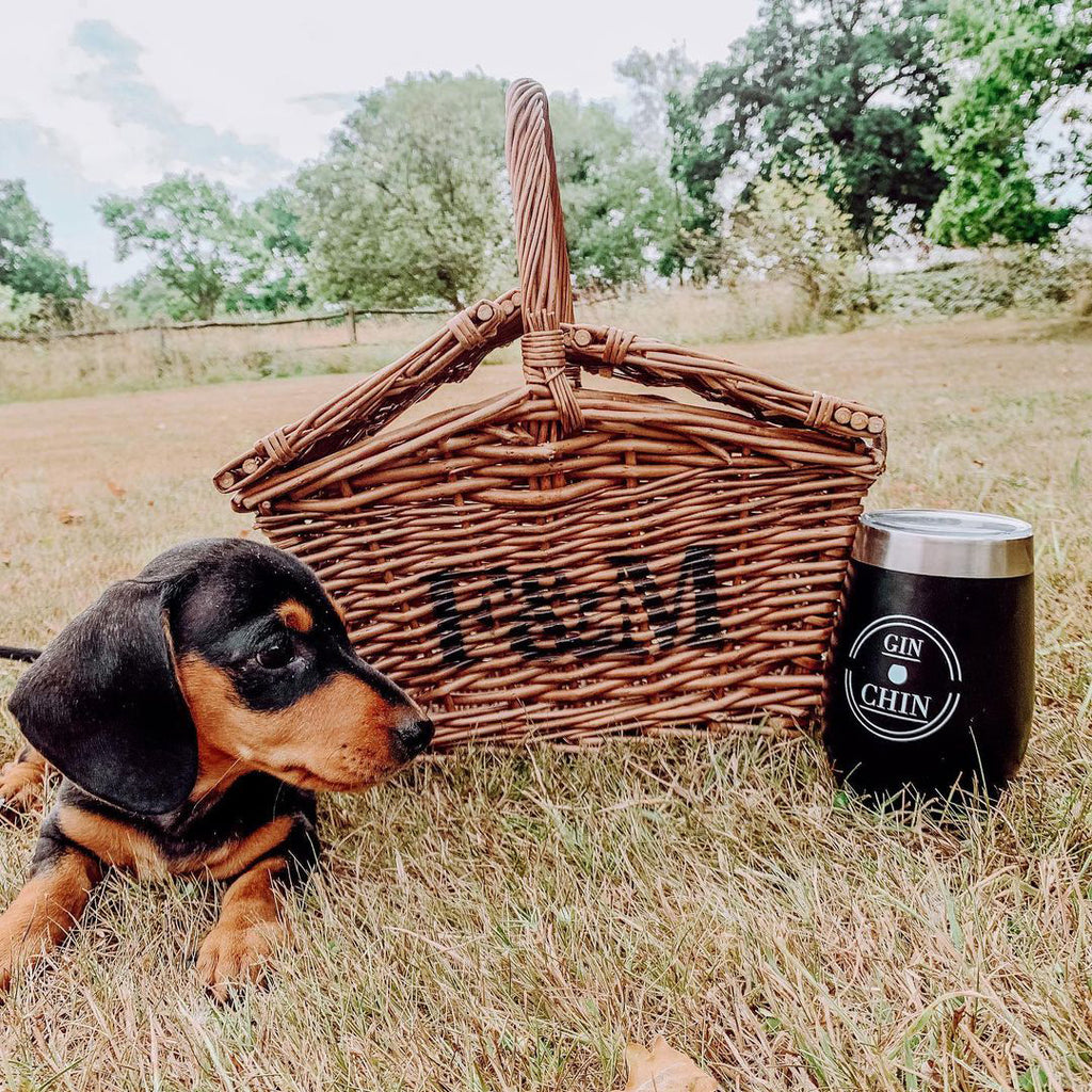 Dog next to a wicker picnic basket and 'GIN & CHIN' insulated tumbler in a grassy outdoor setting