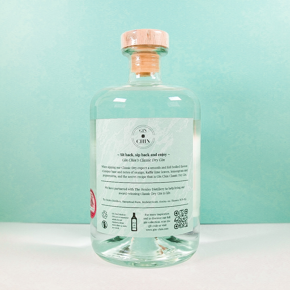 Clear glass bottle of Gin Chin Classic Dry Artisan Gin with a label on a light blue background