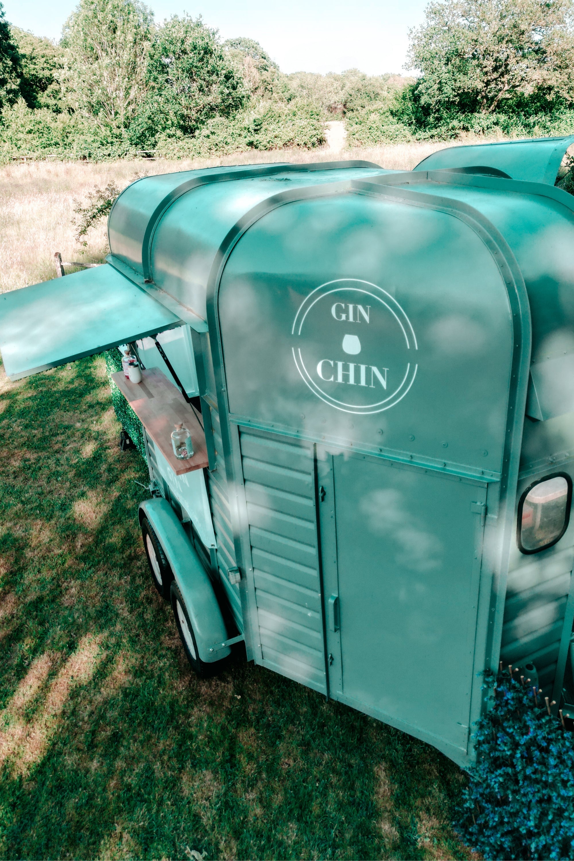 Teal horsebox bar trailer with 'GIN CHIN' branding on a grassy area with trees in the background