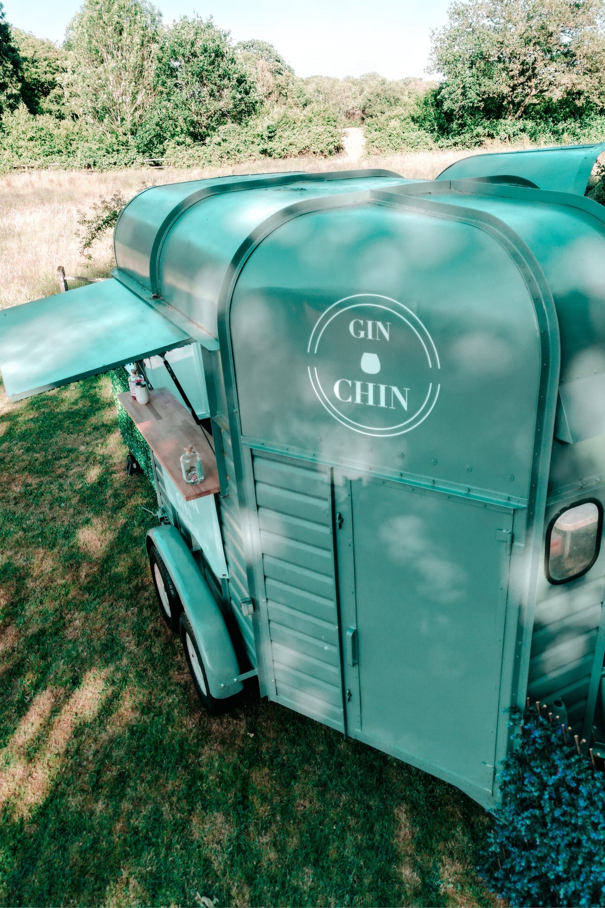 Teal horsebox bar trailer with 'GIN CHIN' branding on a grassy area with trees in the background