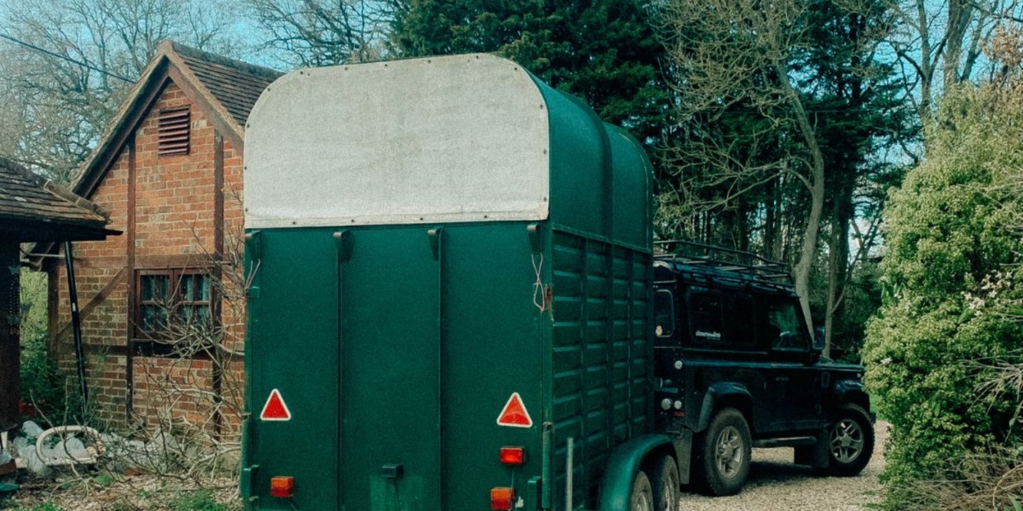 Green horse gin trailer parked next to a brick building with trees in the background