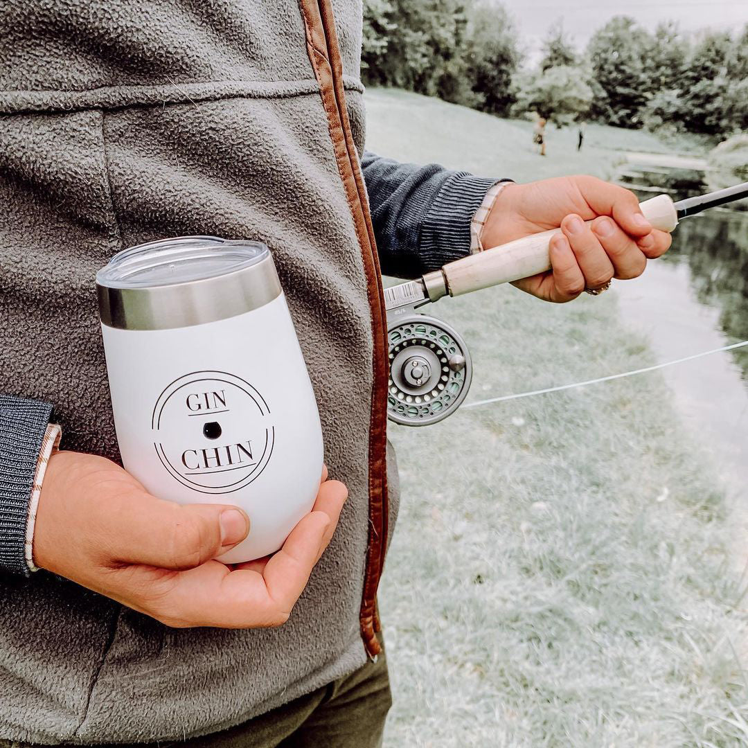 Person holding a white travel tumbler with 'Gin Chin' branding and a fishing rod by a body of water.