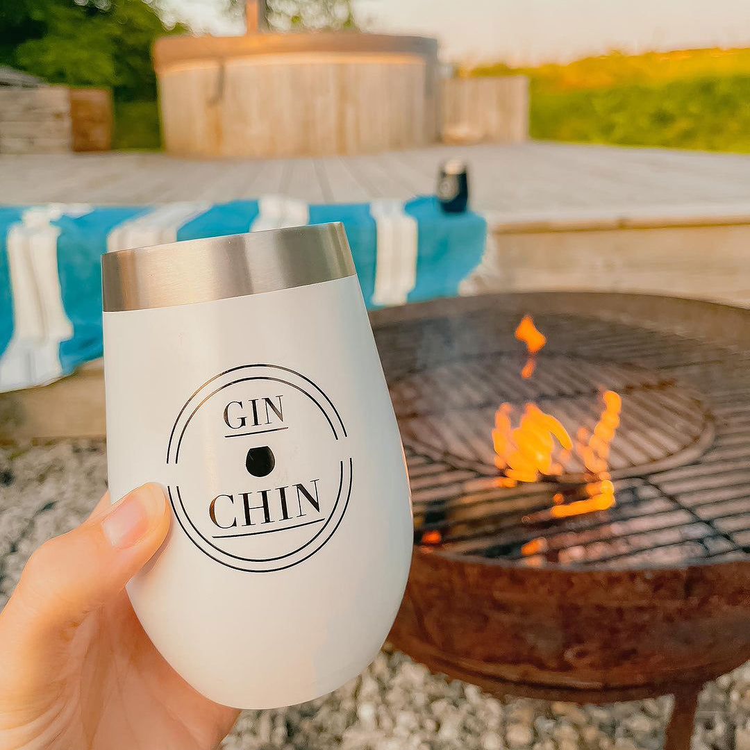 White insulated travel tumbler with 'GIN CHIN' branding held by a hand in front of a fire pit.