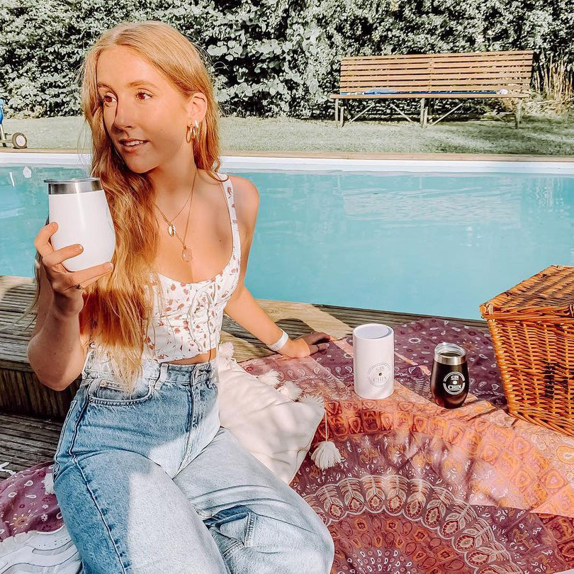 Woman sitting by a pool holding a white insulated tumbler with a picnic setup around her.