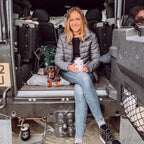 Woman sitting in the back of a vehicle with a dog and a insulated travel cup, smiling at the camera.