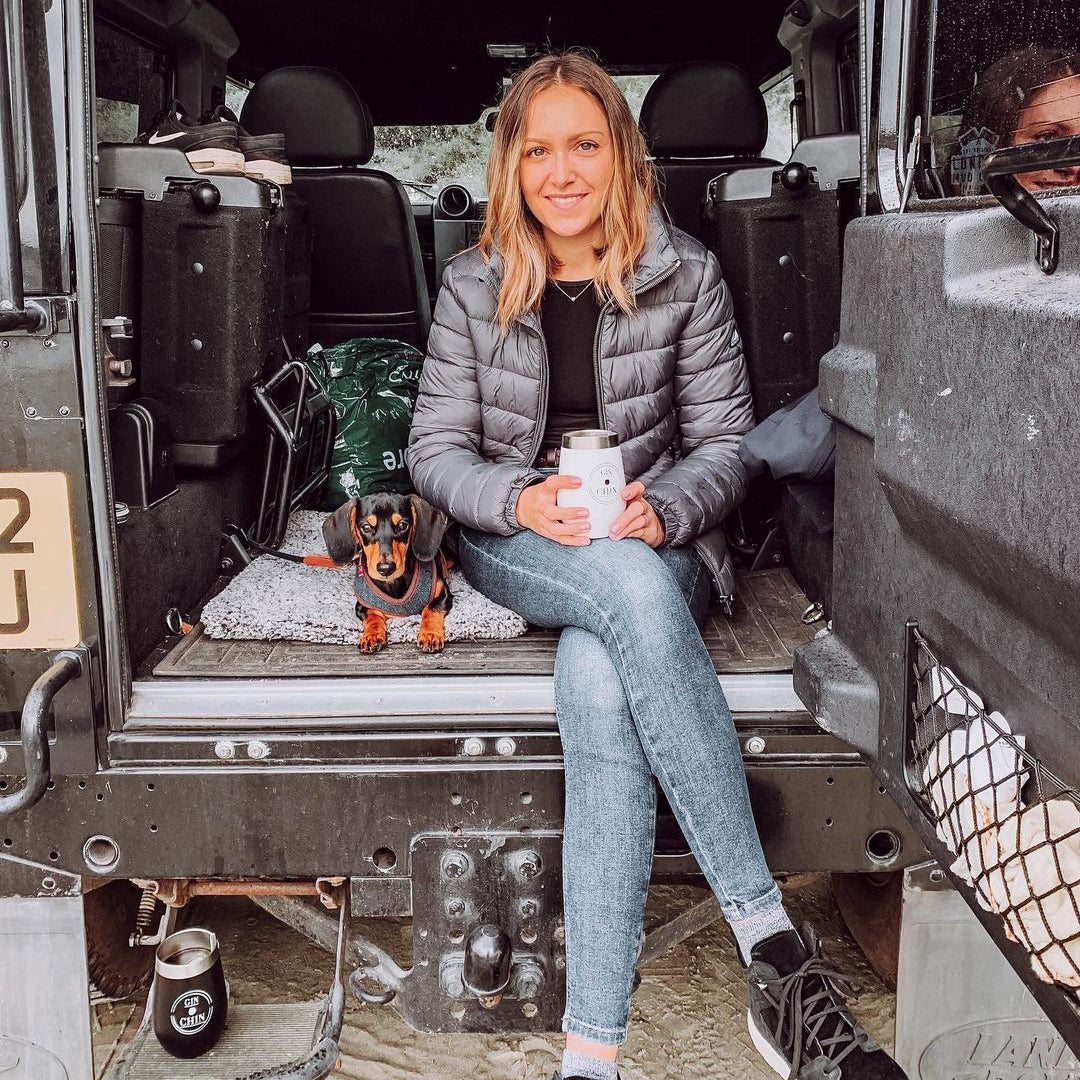 Woman sitting in the back of a vehicle with a dog and a insulated travel cup, smiling at the camera.