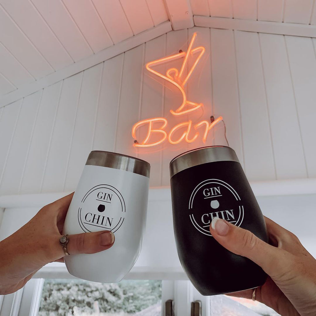Two insulated tumblers labeled 'GIN CHIN' held by hands in front of a neon 'Bar' sign.