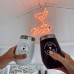 Two insulated tumblers labeled 'GIN CHIN' held by hands in front of a neon 'Bar' sign.