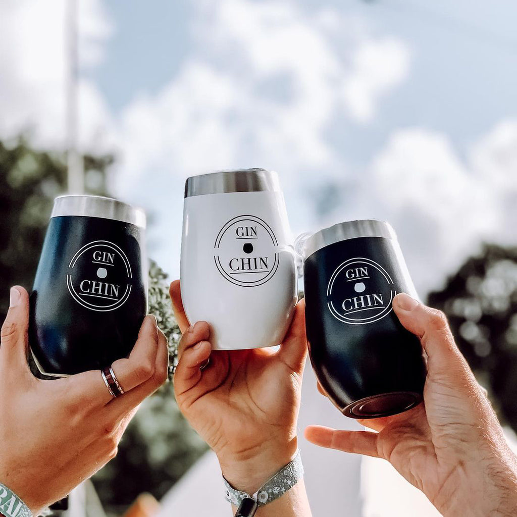 Three black and white 'GIN CHIN' insulated gin tumblers held by hands against a blurred outdoor background.
