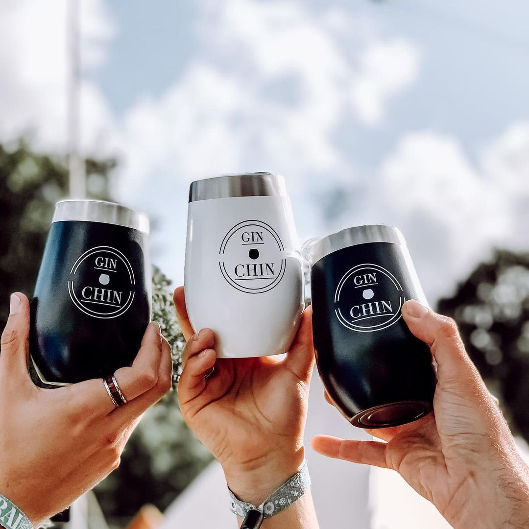 Three black and white 'GIN CHIN' insulated gin tumblers held by hands against a blurred outdoor background.