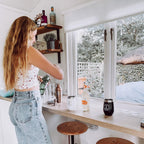 Person standing in a small kitchen with a window view, making a cocktail using a GIN CHIN insulated travel tumbler