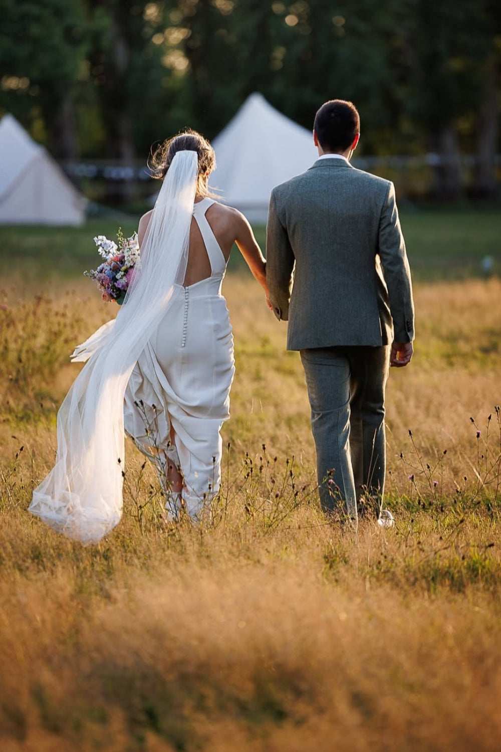 Couple in wedding attire walking hand in hand through a field with tents in the background.