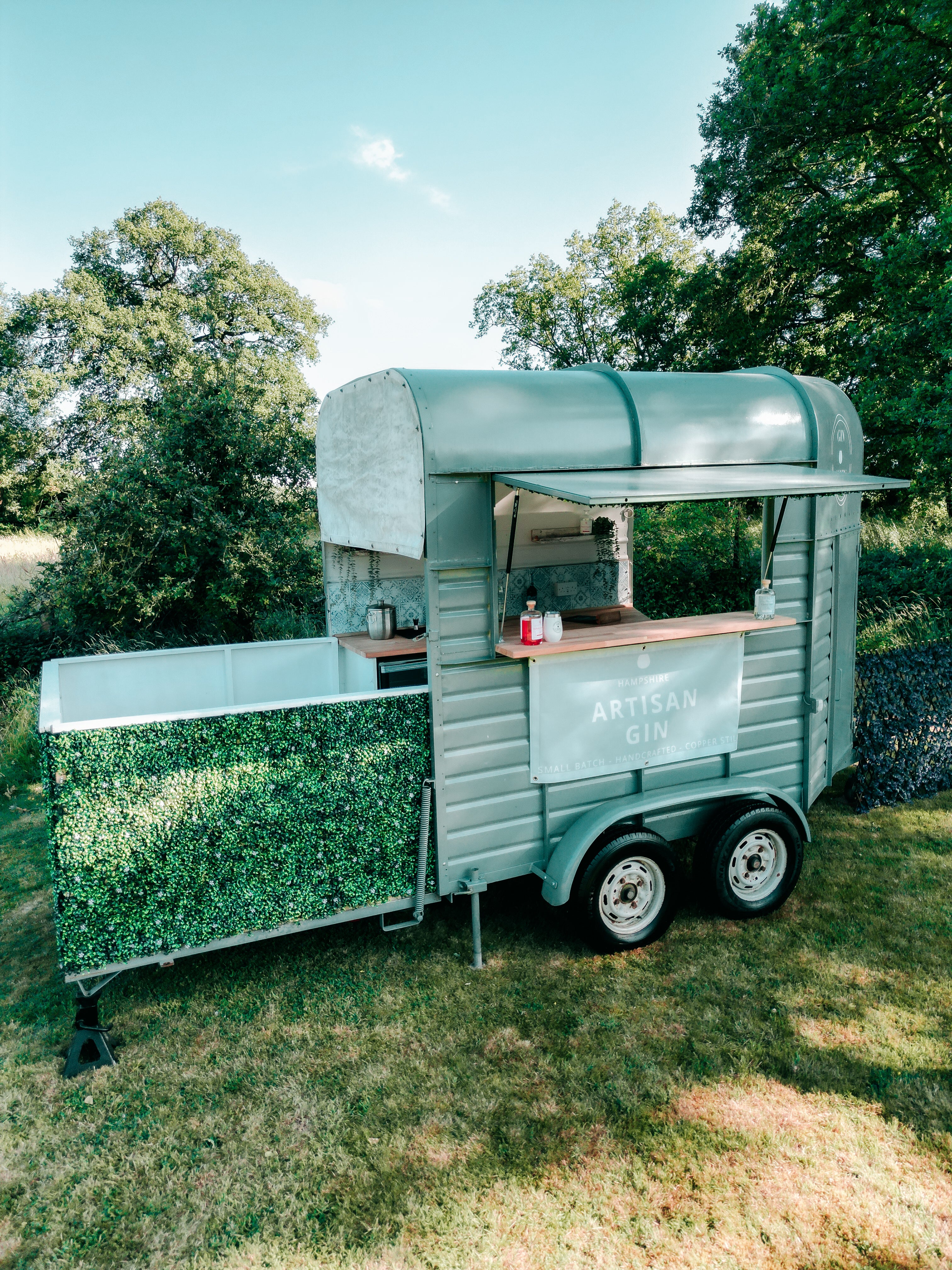 Small mobile gin bar trailer with a green exterior and wooden top, parked outdoors with trees in the background.